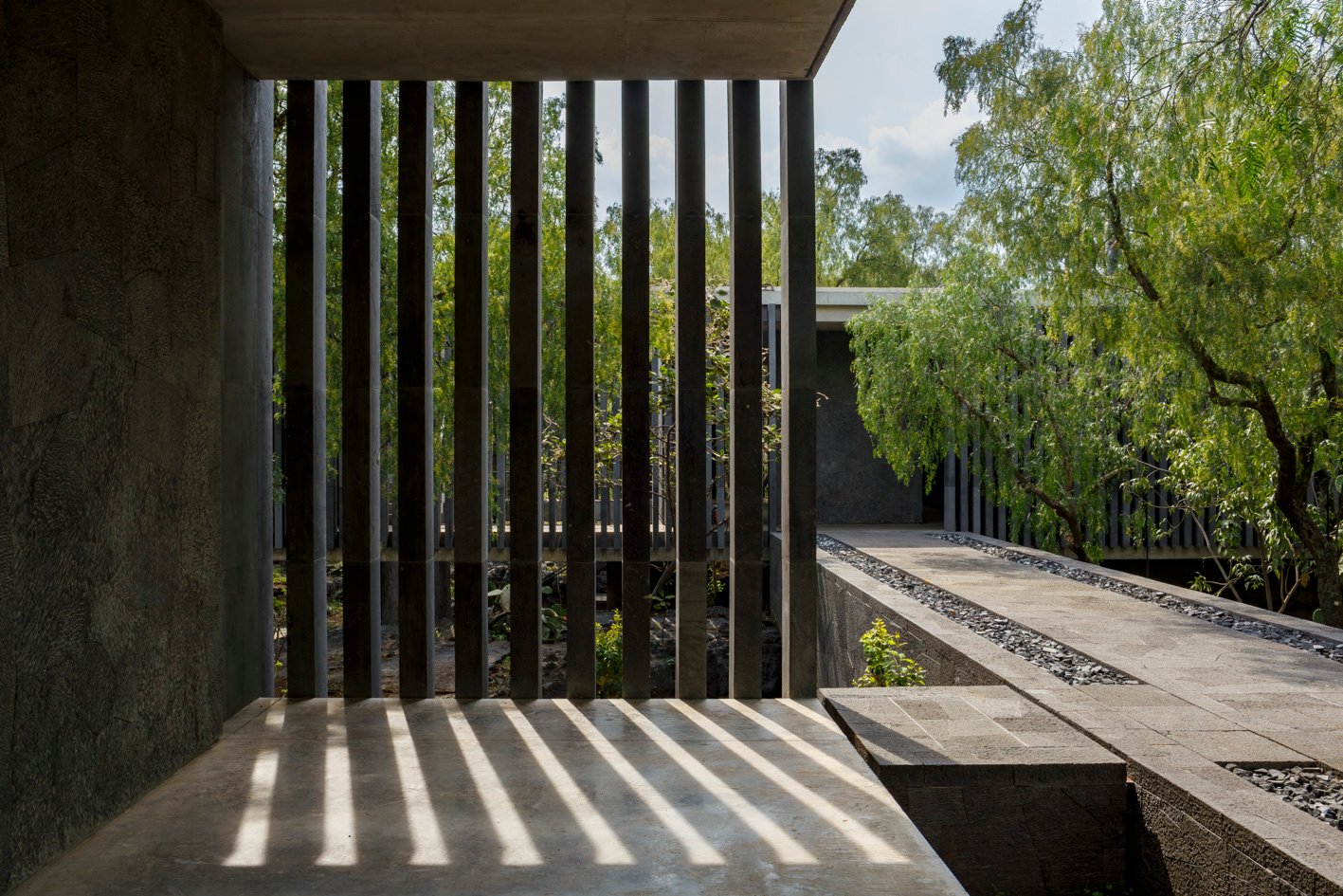 Luz y sombras entre elementos verticales de hormigón en el Museo Anahuacalli, junto a un jardín arbolado.