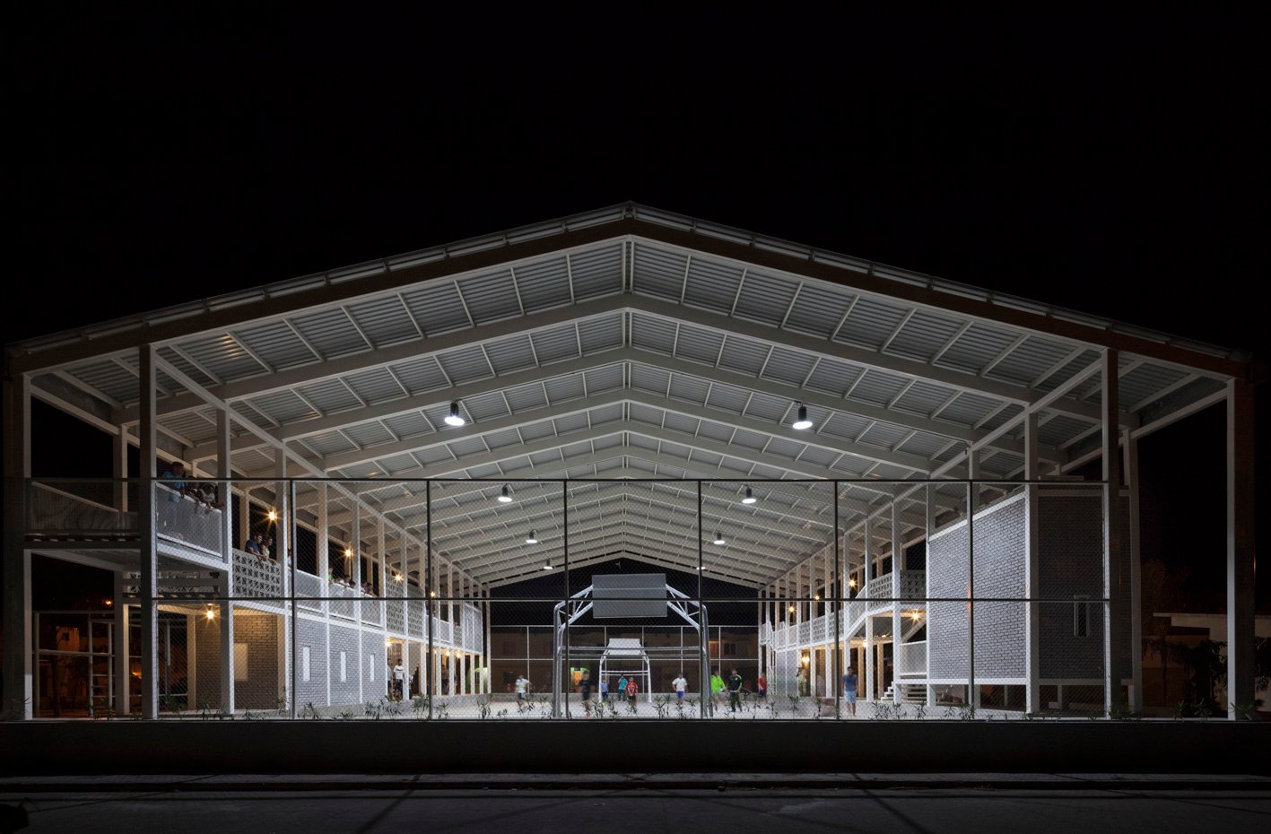 Cancha deportiva cubierta e iluminada de noche, con estructura blanca y personas jugando bajo la cubierta.