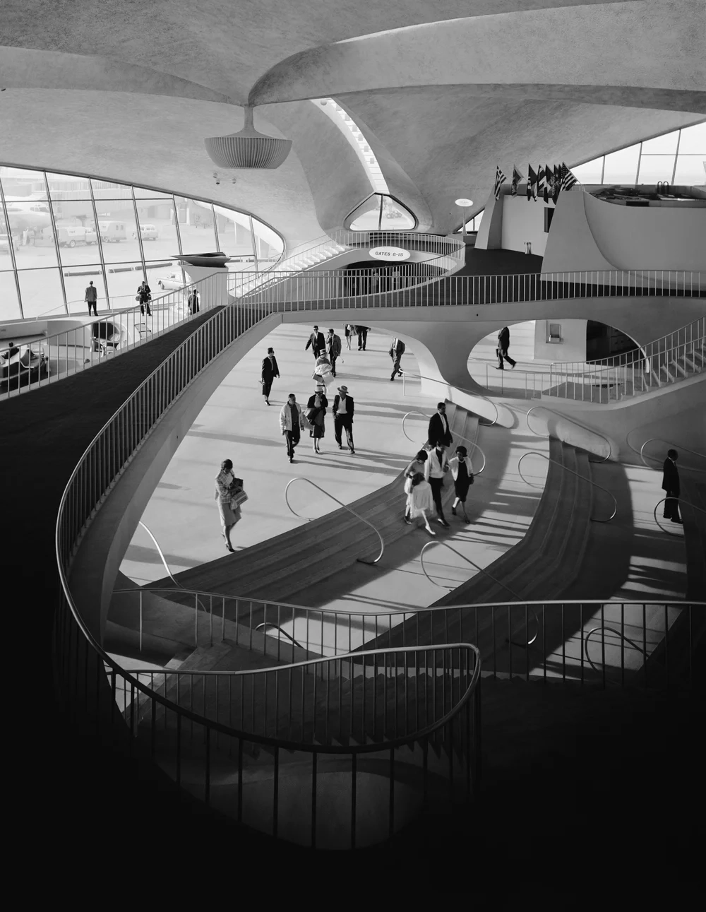 Interior de la terminal TWA en blanco y negro, con pasajeros, escaleras curvas y grandes ventanales.