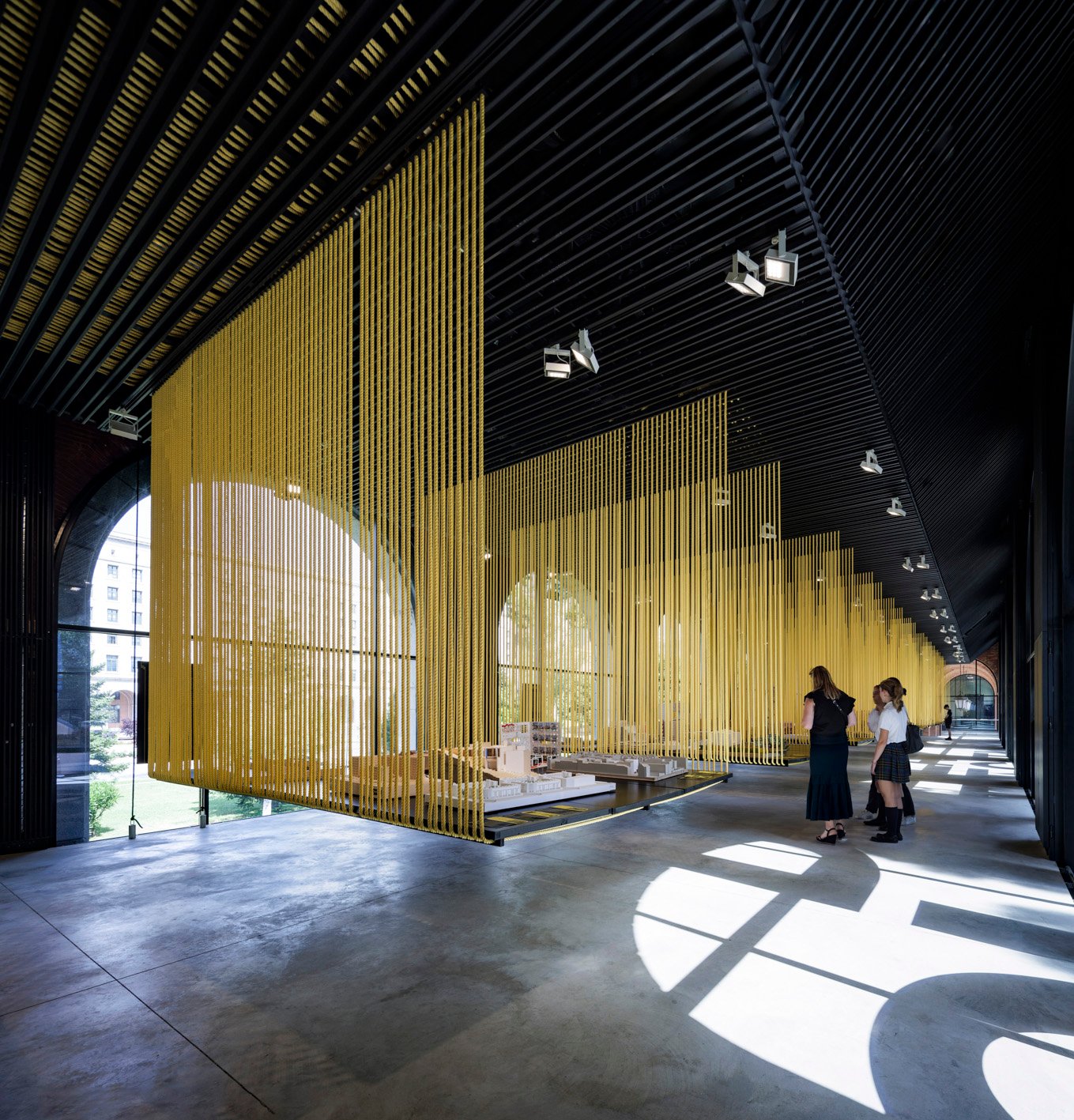 Exposición Habitar España, con maquetas entre cortinas de cuerdas amarillas, visitantes y luz de grandes ventanales.
